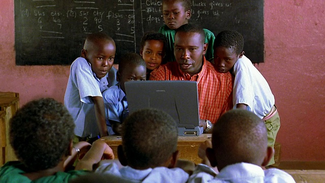 MS向上倾斜，向下倾斜，黑人男子坐在教室里使用电脑，孩子们围着他/肯尼亚视频素材