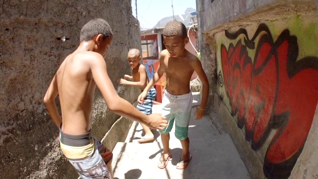 年轻的巴西男孩踢着足球穿过摇摇欲坠的石巷，微笑着对着镜头做着和平手势视频下载