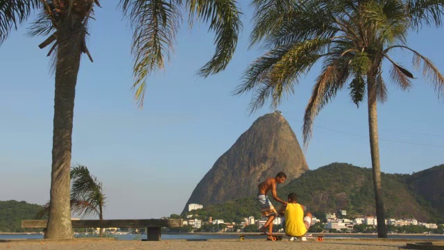 里约热内卢少年滑着滑板来到朋友面前，与朋友握手，坐在滑板上与远处的甜面包山视频素材