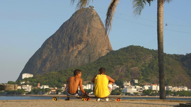 瓜纳巴拉湾(Guanabara Bay)边，里约热内卢青少年坐在滑板上聊天，远处耸立着甜面包(Sugarloaf)视频素材
