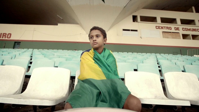 一个小男孩坐在空荡荡的足球场里，身上裹着巴西国旗视频素材