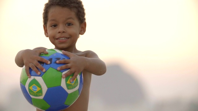 在科帕卡巴纳海滩上，一个小男孩骄傲地拿着巴西足球视频素材