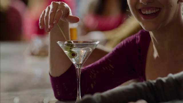 一个女人在马提尼杯里搅拌橄榄，在拥挤的酒吧里大笑视频素材