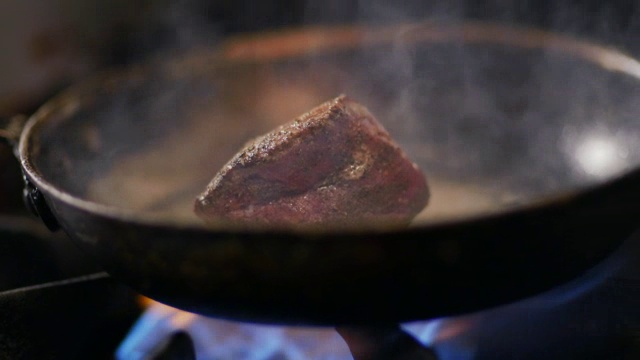 牛排在热气腾腾的铁煎锅里嘶嘶作响，厨师在餐厅厨房里用夹钳翻个身视频素材