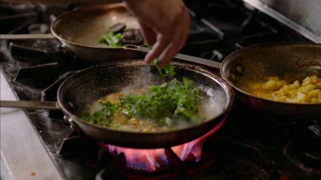 餐厅厨师在厨房里给玉米煮玉米粥拌上绿色蔬菜，翻动着燃烧的煎锅视频素材