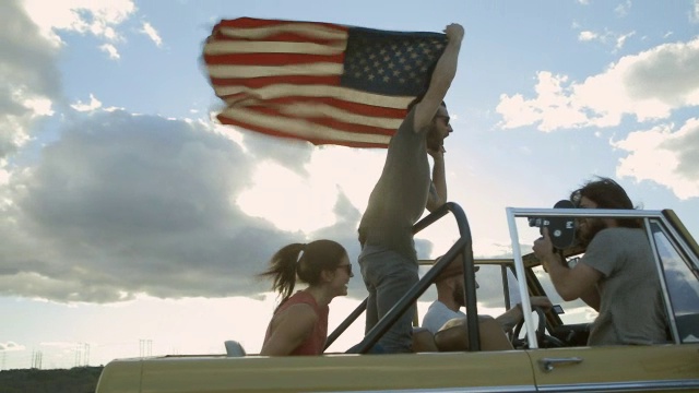 一个年轻人站在野马车的后座上，挥舞着美国国旗，而他的朋友正在用16毫米相机拍摄视频素材