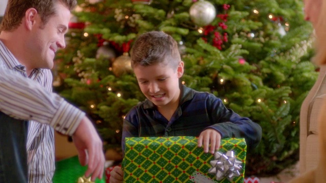 妈妈和爸爸看着男孩在圣诞树下小心翼翼地打开礼物(多莉拍摄)视频素材