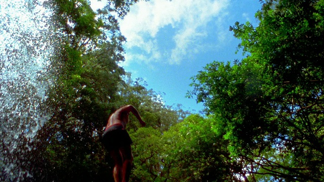 慢动作中拍摄低角度倾斜下的人从瀑布跳进水包围的树木/毛伊岛，夏威夷视频下载