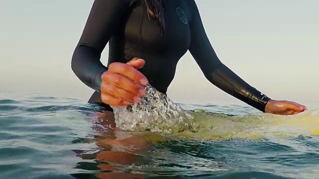 美丽的女性冲浪者坐在冲浪板上，跑步手通过水在日出的冲浪破浪和海浪在大西洋海岸在法国南部。视频下载