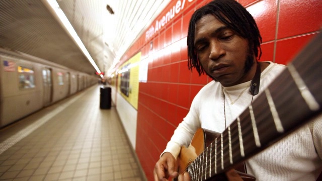 一个靠在红墙上的黑人在地铁站里对着正在行驶的火车弹吉他视频素材