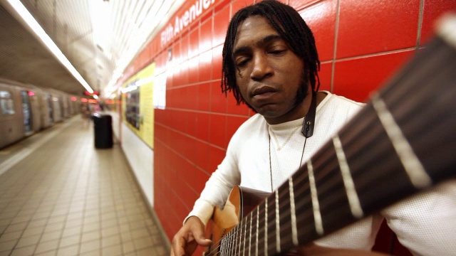 一名黑人靠在红墙上弹吉他，地铁站里行人走在月台上视频素材