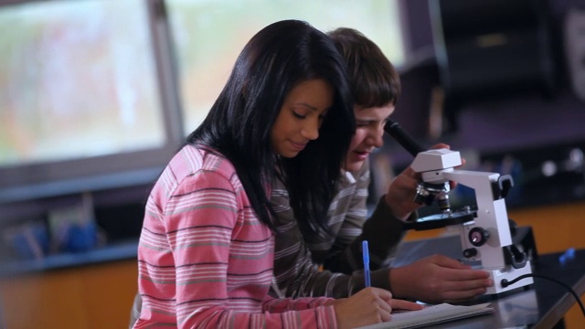 两个学生共用一个显微镜，男学生在显微镜里同行，女学生做笔记视频素材