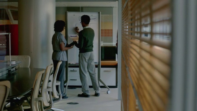 男女商业伙伴在白板上写字，两位时髦的商人一边走在办公室走廊上，一边用平板电脑讨论战略(摄影)视频素材