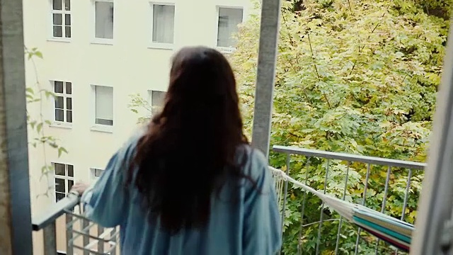 美丽的年轻女子赤脚喝咖啡，穿着超大的牛仔衬衫在阳台上典型的老公寓，德国柏林。视频素材