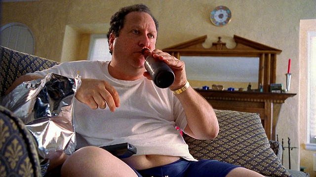 一名穿着t恤的中杯超重男子坐在沙发上，吃着爆米花，喝着啤酒，用着遥控器视频下载