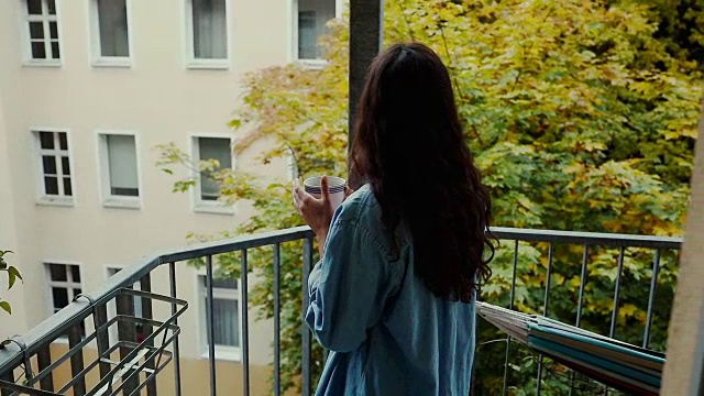 美丽的年轻女子赤脚喝咖啡，穿着超大的牛仔衬衫在阳台上典型的老公寓，德国柏林。视频素材