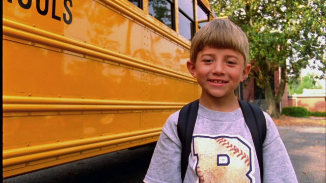 一个小男孩在校车前拿着一个苹果微笑着。视频素材