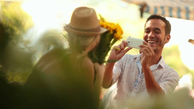 在阳光明媚的市场里，可爱的女孩把脸藏在一束花后，偷看着用智能手机拍照片视频素材