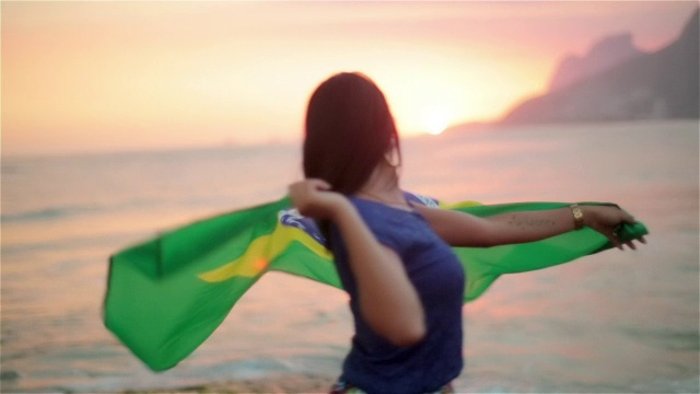 日落时分，伊帕内玛海滩上的女孩在风中舞动巴西国旗视频素材