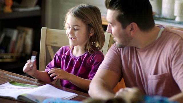 可爱的小女孩轻轻拍了拍爸爸的肩膀，给他看她在餐桌上的魔笔画。视频素材