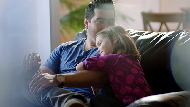 小女孩拥抱和依偎在沙发上的爸爸，他亲吻她的前额。视频素材