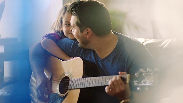年轻的父亲为他的女儿弹奏原声吉他，她靠在他的肩膀上微笑着。视频素材
