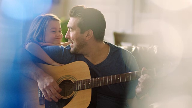 爸爸在家里的客厅里弹奏着原声吉他，女儿则靠在爸爸的肩膀上抚弄着他的头发。视频素材