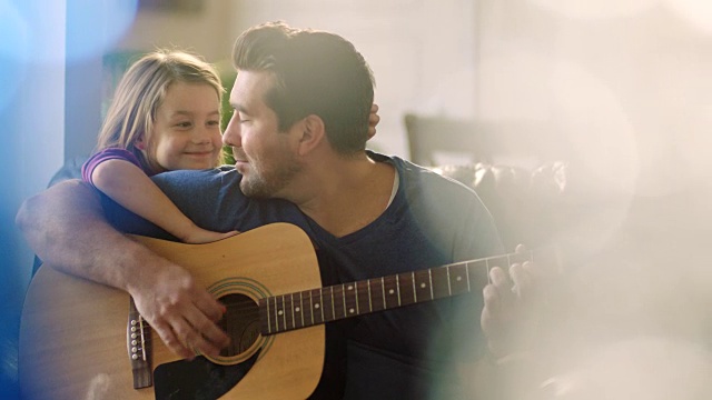 父亲坐在一旁弹奏原声吉他，靠在女儿的肩膀上微笑着看着她。视频素材