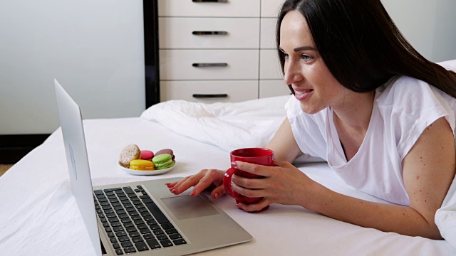 这是一个愉快的女人一边在笔记本电脑上工作一边喝着风味茶的侧面照片视频下载