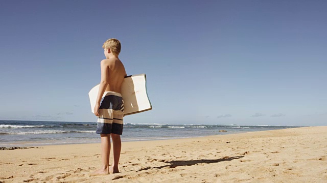小男孩站在沙滩上，拿着冲浪板视频素材