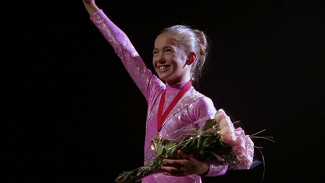 放大中景，年轻的女子体操运动员戴着奖牌，手持鲜花，微笑着挥手视频下载