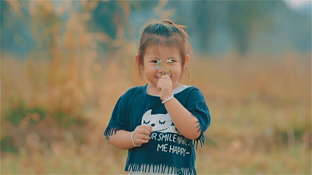 一个微笑的亚洲可爱女孩的肖像看着相机和闻到草地上的花视频素材