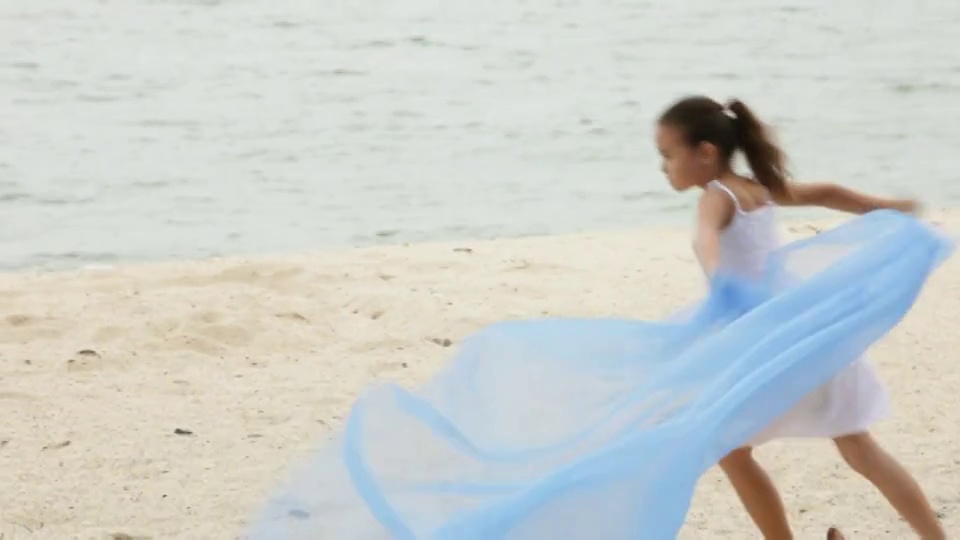 一个小女孩在海滩上跑步，身后挎着一条波浪起伏的蓝色围巾。视频下载