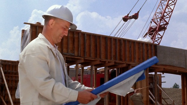 20世纪60年代宽镜头男工人戴着安全帽在建筑工地看蓝图/在背景工作的人视频素材