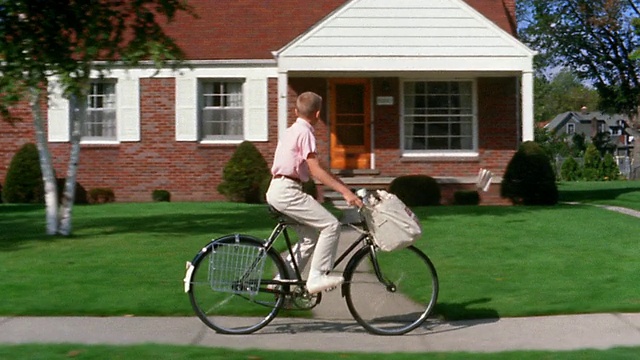 20世纪60年代的宽镜头跟踪拍摄了一个骑着自行车把报纸扔到屋前草坪上的送报纸的男孩视频素材