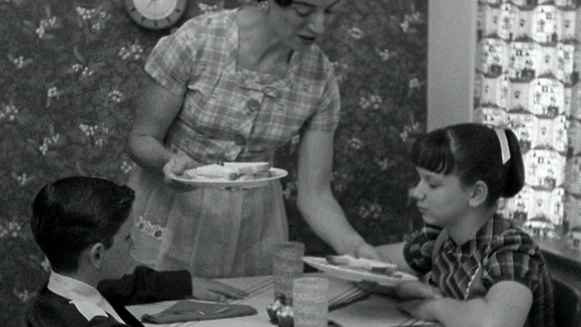 中景再现:一位妇女在厨房的餐桌上为男孩和女孩端上一盘盘三明治视频素材