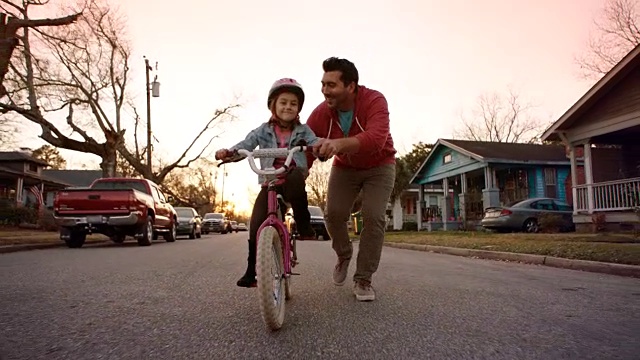 小女孩骑着自行车，对着镜头微笑，爸爸在附近的街道上跑步。视频下载
