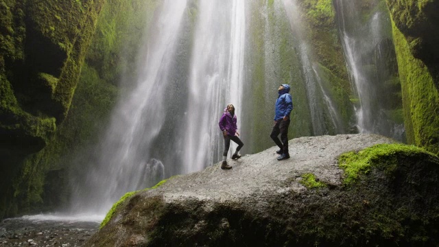 一对夫妇站在瀑布底部的岩石上的宽慢镜头拍摄/冰岛兰加瓦拉斯拉视频下载