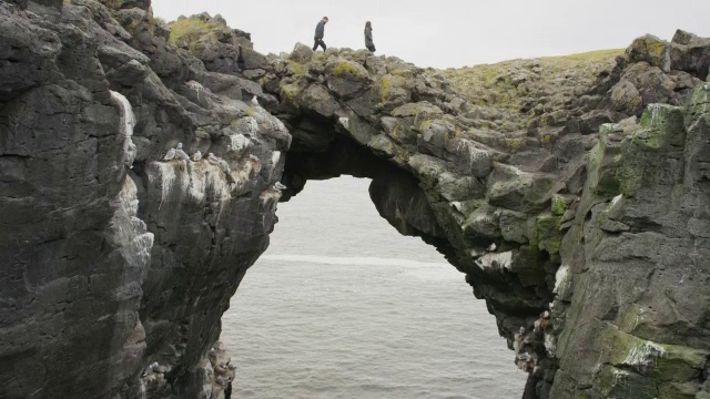 宽吊车拍摄的夫妇行走在拱形岩层上/ Arnarstapi, Snaefellsnes，冰岛视频下载