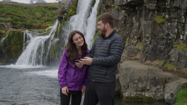在冰岛Snaefellsnes瀑布/ Grundarfjorour附近，一对情侣摆着手机自拍的中景视频素材