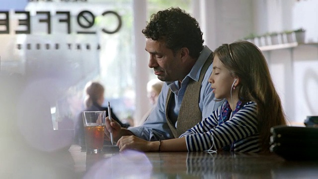 当她的爸爸在当地的一家咖啡店里用平板电脑工作时，小女孩一边听着音乐一边唱歌。视频素材