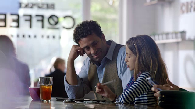 父亲微笑着向女儿展示如何在现代咖啡店里使用平板电脑。视频素材