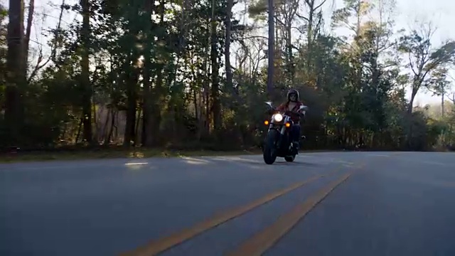 年轻女子骑着摩托车在空旷的高速公路上飞驰，树叶飘落，太阳在树木间闪耀。视频下载