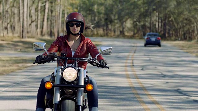 穿着红色皮夹克的年轻女子骑着摩托车在弯弯曲曲的道路上游弋。视频素材