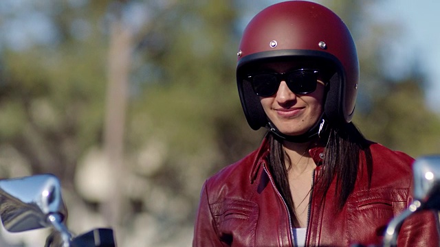 穿着皮夹克和头盔的年轻女子在摩托车上笑，对着镜头微笑。视频下载