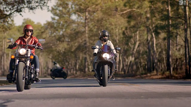 两个骑摩托车的女人在森林公路上行驶，另一个骑摩托车的人经过。视频素材