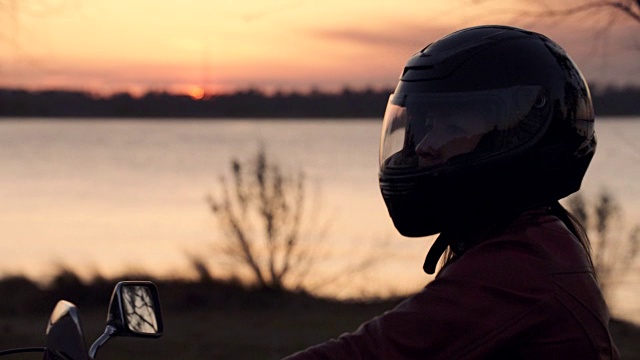 女人放下头盔上的遮阳板，骑上摩托车，太阳在河上落下。视频下载