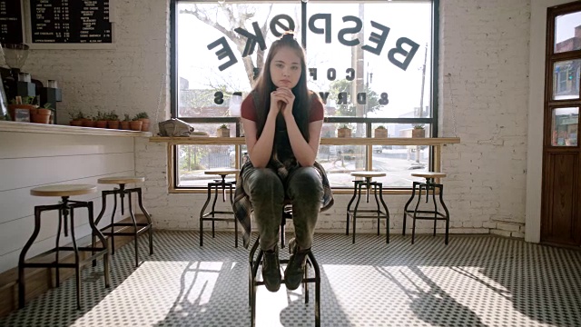 年轻女孩坐在酒吧的凳子上盯着镜头在现代咖啡店。视频素材