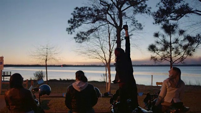 一名女子和朋友坐在摩托车上，站起来向空中挥舞拳头。视频下载