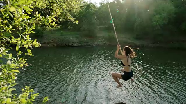 一个女孩从河岸上荡着绳子跳进水里溅起水花。视频素材
